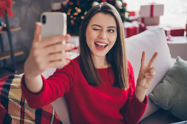 Chica positiva sentarse sofá tomar selfie en el teléfono inteligente hacer v-signo de parpadeo en casa en el interior con adorno de Navidad de x-mas —  Fotos de Stock