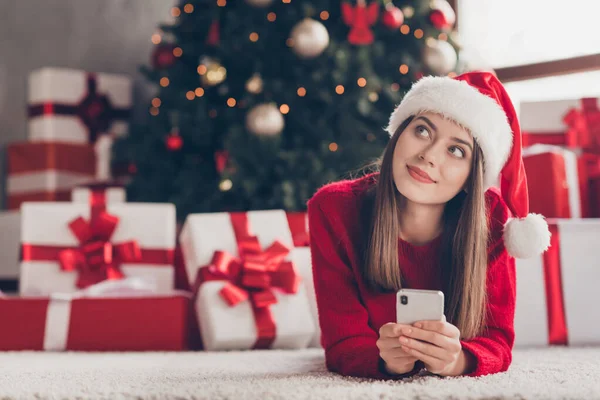 Joven señora bonita mentira piso mantenga móvil buscar desgaste santa gorra jersey rojo en decorado x-mas sala de estar en el interior — Foto de Stock