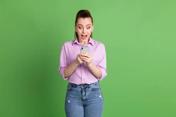 兴奋的女孩用智能手机给人留下深刻印象的照片就像社交媒体的通知，令人印象深刻的尖叫，穿着紫色斜纹棉布牛仔裤，与绿色背景隔离 — 图库照片