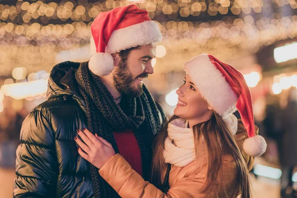 Foto de casal afetuoso abraçando sob a noite ao ar livre x-mas luzes desgaste santa claus cap inverno temporada cachecol — Fotografia de Stock