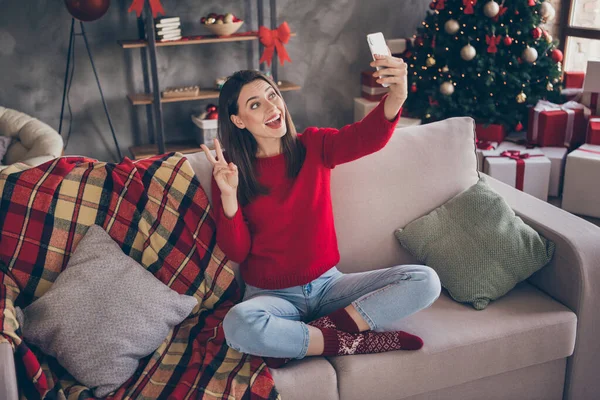 Foto de longitud completa de chica sentarse sofá tomar selfie en el teléfono inteligente hacer v-signo en la casa en el interior con la decoración de Navidad de x-mas —  Fotos de Stock