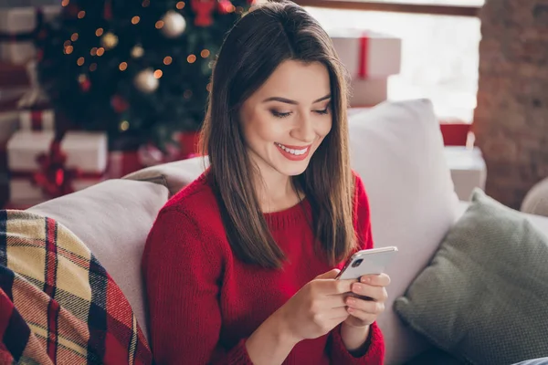 Фото позитивної дівчини, що друкує лист на смартфоні, сидять затишний диван в будинку з різдвяним орнаментом на Різдво — стокове фото