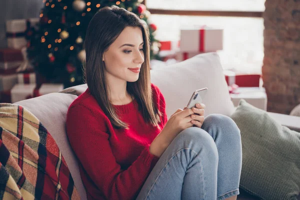 Фото девушки сидеть диван читать рождественские новости на смартфоне носить красный свитер в доме в помещении с рождественским украшением — стоковое фото
