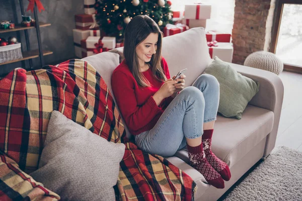 Pełny rozmiar zdjęcie pozytywnej dziewczyny czat na smartfonie siedzieć kanapa nosić czerwony sweter w domu w domu z x-mas Boże Narodzenie noel ozdoba — Zdjęcie stockowe