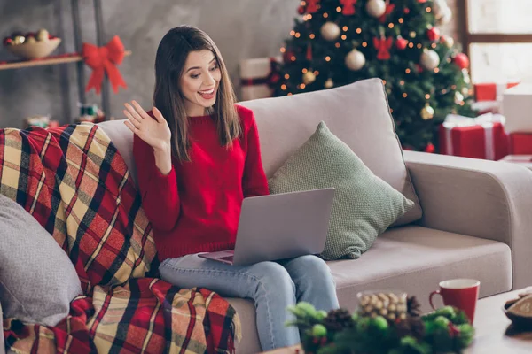 Foto de la joven señora bonita sentarse sofá mantenga la computadora agitando el brazo usar pantalones vaqueros jersey rojo en decorado x-mas sala de estar casa interior —  Fotos de Stock