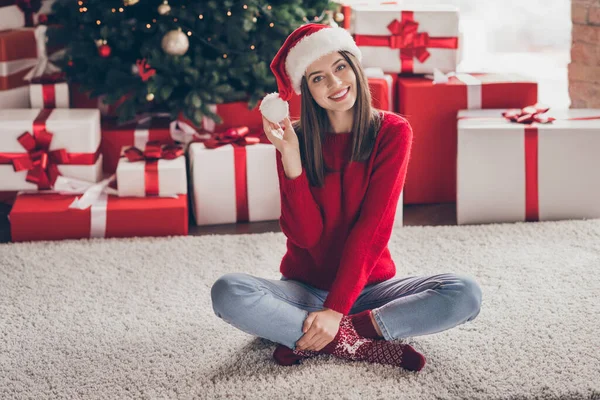 Fotografie krásné dámy sedět koberec dárkové krabice hrát pompon nosit santa headwear červený svetr ponožky džíny v zdobené x-mas obývací pokoj uvnitř — Stock fotografie