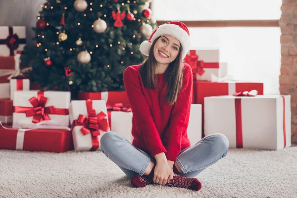 Fénykép a csinos fiatal lány ajándék dobozok karok tartani keresztbe lábak viselnek Mikulás fejfedő piros pulóver díszített x-mas nappali beltérben — Stock Fotó