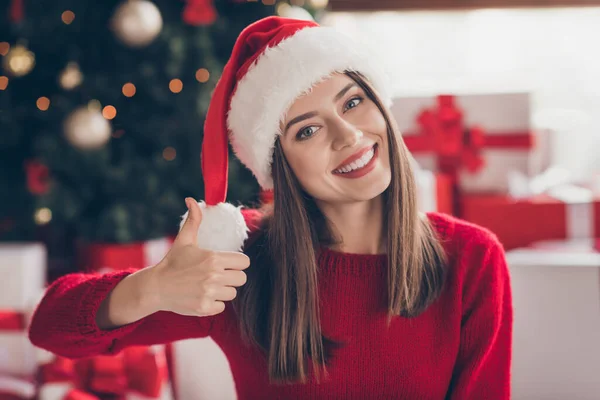 Güzel bir kadın elini kaldırır, başparmağını kaldırır dişlek bir gülümseme takar, Noel Baba 'nın başlığını giyer, Noel Baba' nın kırmızı süveterini giyer, kapalı bir oturma odasında Noel Baba süveteri giyer. — Stok fotoğraf