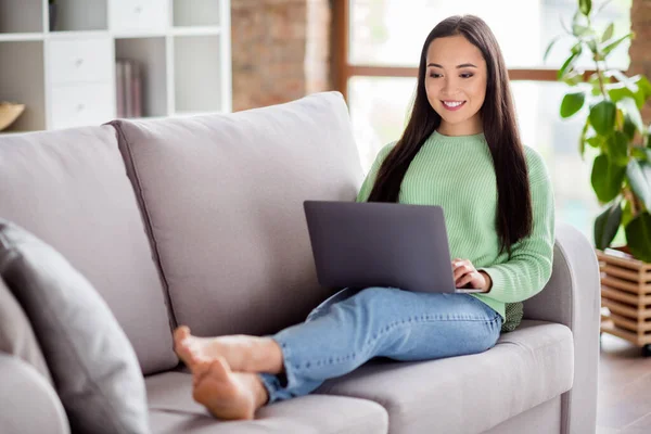 Foto de tamaño completo de chica coreana positiva estudiante de la universidad estudio portátil de trabajo remoto preparar el proyecto de desgaste pantalones vaqueros suéter verde pies desnudos se encuentran diván en casa en el interior — Foto de Stock