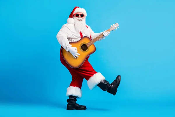 Plná velikost fotografie bláznivý cool Santa Claus s velkým břichem vousy hrát na kytaru x-mas vánoční výkon nosit sluneční brýle podvazky montérky izolované přes modré barvy pozadí — Stock fotografie