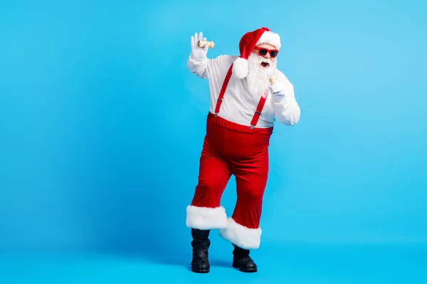 Teljes test fotó stílusos őrült funky hűvös hipster santa claus nagy has énekelni x-mas karácsonyi dal bors mikrofon kopás piros nadrágtartó overál napszemüveg elszigetelt kék szín háttér — Stock Fotó