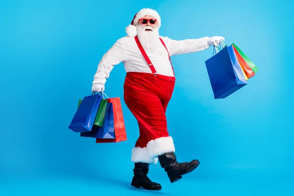 Full längd profil sida foto av galna moderna vit grått hår skäggig tomte gå shopping köpa jul jul påsar bära röda hängslen overaller stövlar isolerad blå färg bakgrund — Stockfoto
