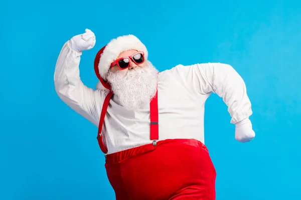 Bild av gammal man grått skägg dans höja handen rolig retro flytta bära Santa claus x-mas kostym mössa huvudbonad solglasögon hängslen vit skjorta handskar isolerad blå färg bakgrund — Stockfoto