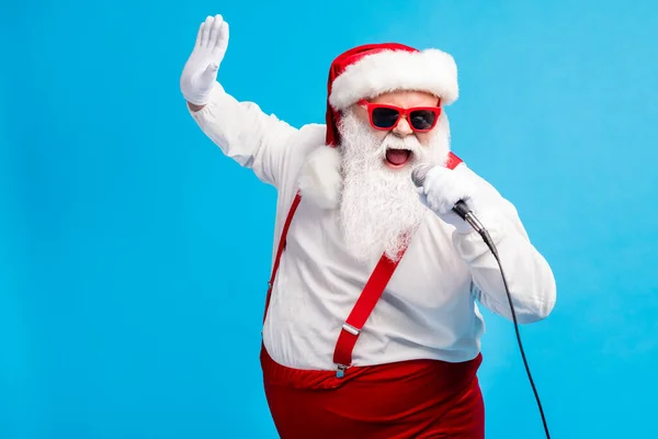 Photo of old man grey beard hold mic open mouth sing karaoke raise arm wear santa claus x-mas costume cap sunglass suspenders white shirt gloves isolated blue color background — Stock Photo, Image