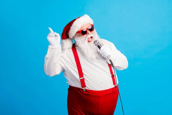 Foto di pazzo moderno Babbo Natale con grande pancia barba cantare microfono x-mas Natale canzone indossare stile bretelle alla moda berretto tuta isolata su colore blu — Foto Stock