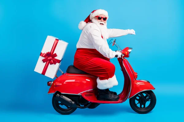 Full size profiel zijkant foto van stijlvolle moderne witte grijze haar bebaarde kerstman rijden motor fiets leveren x-mas kerst tijd geschenkdoos dragen rood kostuum geïsoleerde blauwe kleur achtergrond — Stockfoto
