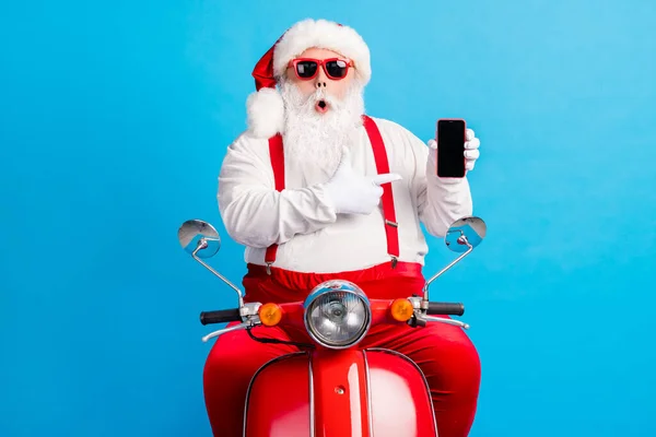 Foto van verbaasd grijs wit haar bebaarde kerstman x-mas kerst scooter bestuurder wijzen vinger smartphone dragen zonneglas overall bretels geïsoleerde blauwe kleur achtergrond — Stockfoto