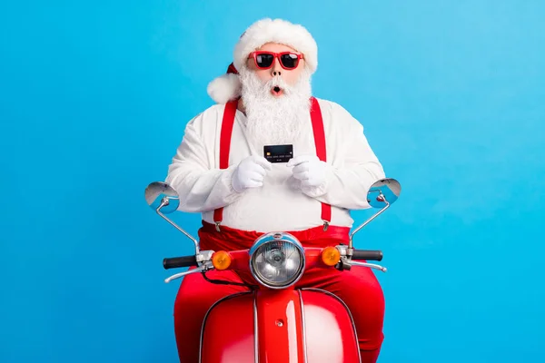 Foto van moderne gekke verbaasd santa claus met grijze baard rijden rode x-mas kerst scooter houden credit card dragen bretels overalls hoofddeksels geïsoleerde blauwe kleur achtergrond — Stockfoto