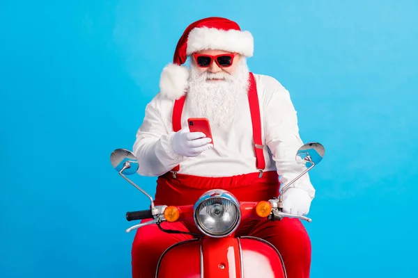 Foto van stijlvolle witte grijze haar bebaarde santa claus drive scooter typen x-mas kerst groet smartphone dragen rode partij bretels overalls cap geïsoleerd over blauwe kleur achtergrond — Stockfoto