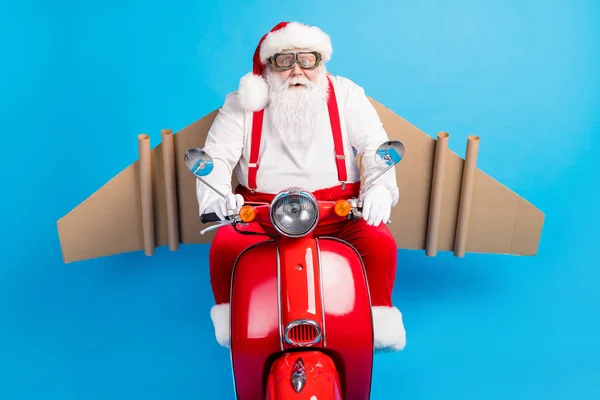 Foto van grappige gekke kerstman rijden scooter snelle snelheid met karton ambachtelijke vleugels vliegen in de lucht dragen moderne stijlvolle bretels overalls bril bril kap geïsoleerde blauwe kleur achtergrond — Stockfoto