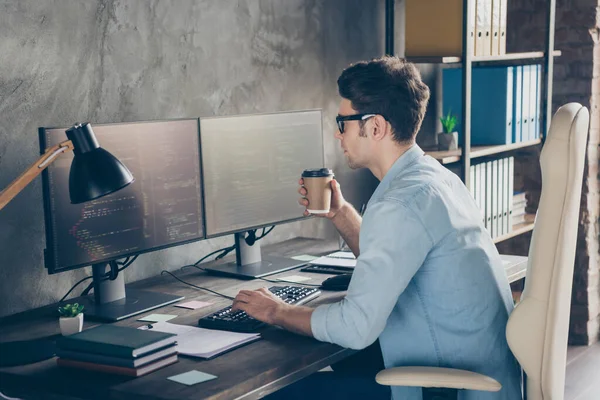 Фотографія з боку профілю адміністратора зосередженого хлопця має багато тестування комп'ютерних робочих столів для роботи на екрані стола для сидіння, що тримає кавовий напій на робочому місці — стокове фото