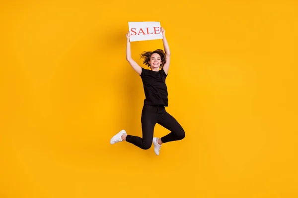Вид в полный рост на прекрасную тонкую жизнерадостную девушку, прыгающую, держа в руках открытку со словом продажа, изолированный ярко-желтым цветом фона — стоковое фото