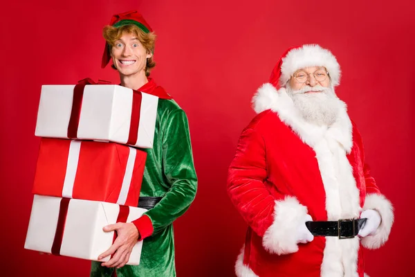 Foto di due uomini Babbo Natale palme cintura elfo tenere molte scatole nastro usura x-mas costume cappotto cintura tappo occhiali isolato colore rosso sfondo — Foto Stock