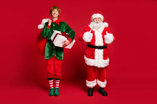 Full length photo of two men santa elf hold bag box raise thumb up wear x-mas costume coat cap glasses boots isolated red color background — Stock Photo, Image
