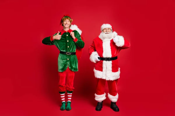 Полная длина фото двух человек Санта-эльф держать большой мешок поднять большой палец вверх износа костюма ботинки колпак ботинки изолированные красный цвет фона — стоковое фото