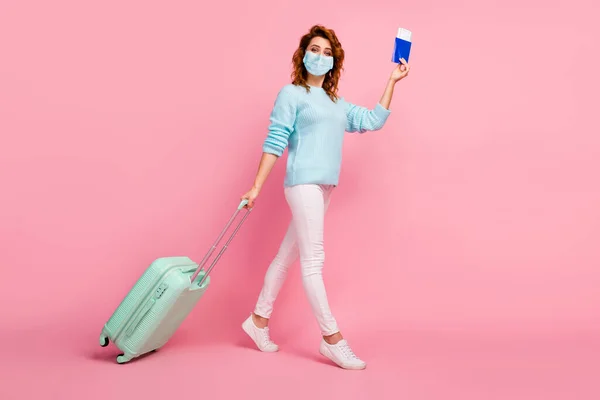 Visualizzazione completa di bella ragazza dai capelli ondulati a piedi portando bagagli passaporto indossare maschera isolata su sfondo rosa pastello colore — Foto Stock