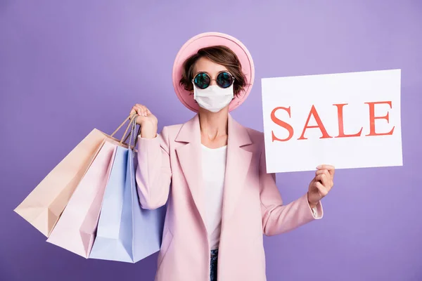 Portret van jonge dame houden aankopen te koop banner dragen griep veilig masker bril roze cap jas geïsoleerd op paarse achtergrond — Stockfoto