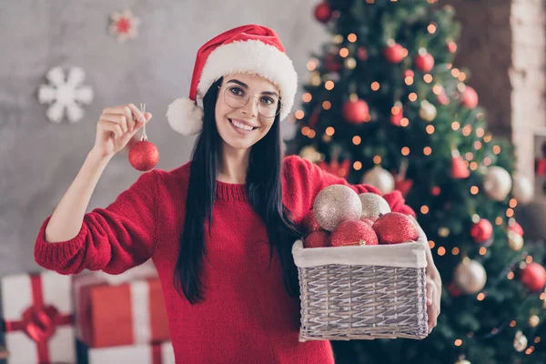 Φωτογραφία του θετικού κοριτσιού φορούν santa claus headwear κρατήσει Χριστούγεννα μπάλες κουτί στο σπίτι με χριστουγεννιάτικα στολίδι πνεύμα — Φωτογραφία Αρχείου