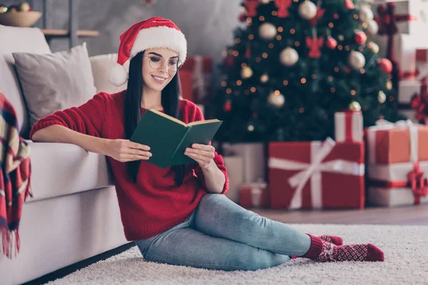 Kızın tüm vücudu Noel Baba 'nın başlığını takıyor. Oturma halısı takıyor. Evde kitap okuyor ve Noel süsleri takıyor. — Stok fotoğraf