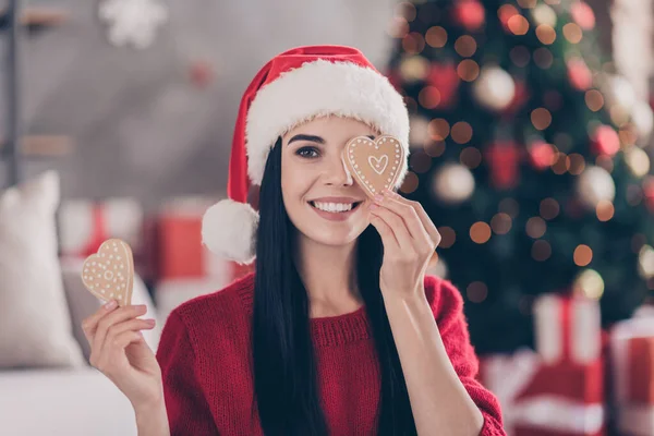 Hoş bir kadının fotoğrafı. İki kalp figürlü bisküvi, göz kapağı, kapalı oturma odasında Noel Baba şapkası kırmızı süveter. — Stok fotoğraf