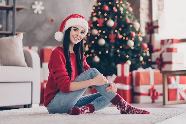 Фото молодої милої дівчини сидить килим обіймає ногу виглядає камера носити Санта-кап червоний светр джинсові шкарпетки в декорованій вітальні в приміщенні — стокове фото