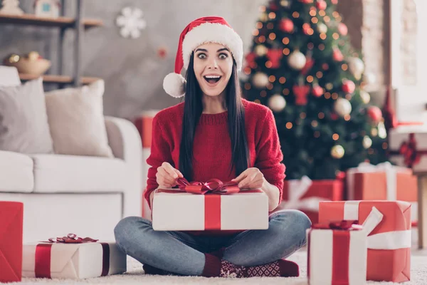 Фото юной леди сидеть на полу держать unboxing подарок открыть рот носить Санта шапка красный пуловер джинсы носки в украшенной гостиной в помещении — стоковое фото