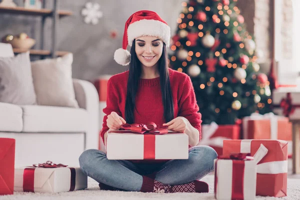 Foto van een charmant meisje zit vloer hold uitpakken gift handen lint dragen santa hoofddeksel rood pullover jeans sokken in ingerichte woonkamer binnen — Stockfoto