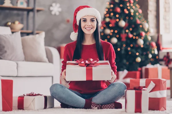 Foto av söt ung flicka sitta golvet hålla stora presentförpackning bära Santa huvudbonader röd tröja jeans strumpor i dekorerat vardagsrum inomhus — Stockfoto