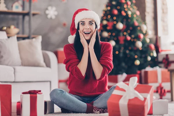Foto av söt ung flicka sitta golv armar kindben öppen mun bära Santa cap röd pullover i dekorerat vardagsrum inomhus — Stockfoto