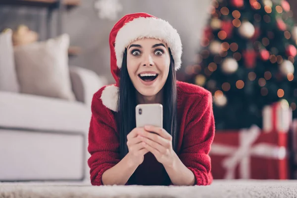 Фото шокированной леди лежал пол держать смартфон открытым ртом носить Санта шапка красный свитер в украшенной гостиной в помещении — стоковое фото