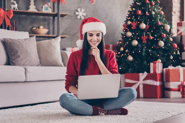 Foto der netten Dame sitzen Teppich halten Netbook Hand Kinn tragen Weihnachtsmannmütze roten Pullover Jeans Socken in dekorierten Wohnzimmer drinnen — Stockfoto
