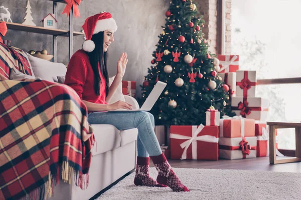 소파에 앉아 있는 젊은 여성의 사진이 인터넷으로 손을 흔들고 있습니다. 손에는 산타 모자에 붉은 스웨터를 입고 실내 실내 실내 장식을 하고 있습니다. — 스톡 사진