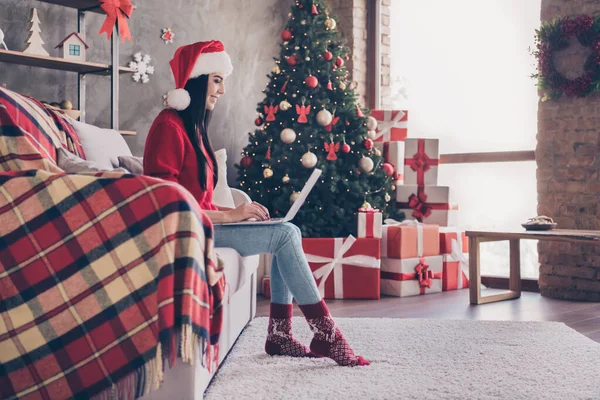 Foto av charmig ung dam sitta soffa hålla netbook bära Santa cap röd tröja denim jeans strumpor i dekorerat vardagsrum inomhus — Stockfoto