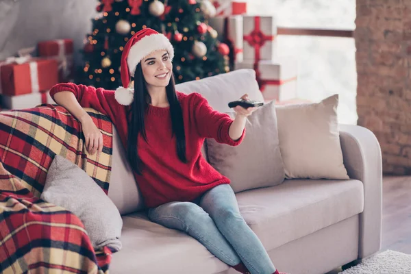 Photo of girl sit sofa hold remote control toothy smile wear santa headwear red sweater jeans in decorated living room indoors