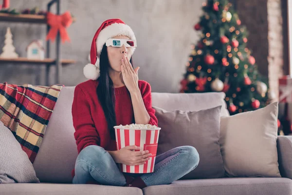 Photo of young lady sit sofa hold pop corn paper box palm close mouth wear 3d glasses santa hat red sweater in decorated living room indoors