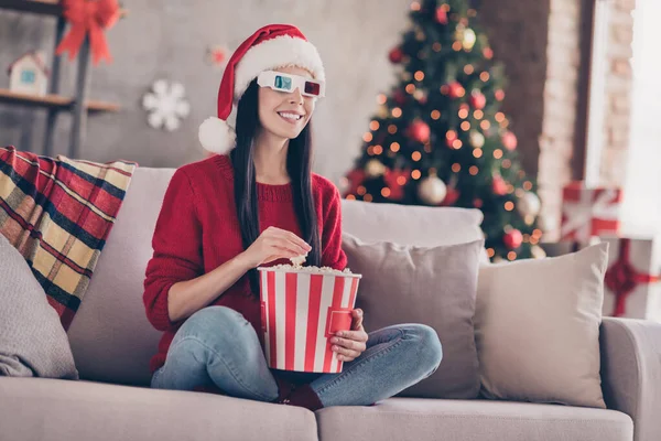 Photo of young girl sit couch hold popcorn paper bucket wear 3d glasses santa cap red pullover jeans in decorated living room indoors — Stock Photo, Image