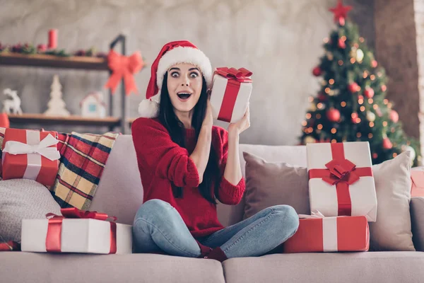 Güzel bir kızın fotoğrafı şaşkın şaşkın oturur yüzü açık kanepede oturur hediye kutusu takar Noel Baba şapkası kırmızı kazak giyer kapalı salonda süslenmiş kot pantolon giyer — Stok fotoğraf