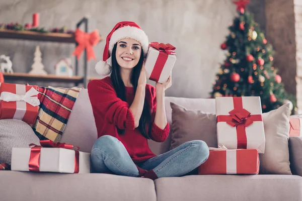 Фото очаровательной девушки сидеть диван держать настоящее время догадаться носить Санта головной убор красный свитер джинсы в украшенной гостиной в помещении — стоковое фото