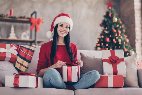 Güzel bir kızın fotoğrafı kanepeye otur boks hediyesi kurdelesini aç birçok hediye Noel Baba şapkası kırmızı kazak kotunu iç mekanda dekore edilmiş salonda giy — Stok fotoğraf