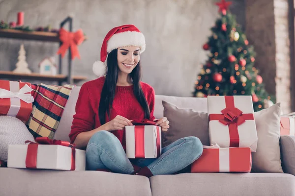 소파에 앉아 있는 예쁜 소녀의 사진 포장을 풀지 않은 경품 상자를 들고 있는 많은 선물들은 실내 거실에 장식 된 산타 모자의 붉은 스웨터를 입고 있다 — 스톡 사진